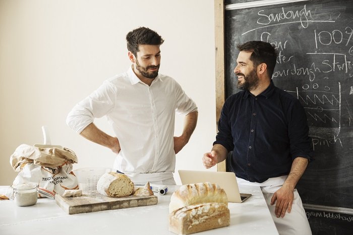 Two bakers standing at a table, using a laptop computer, freshly baked bread, a blackboard on the wall.